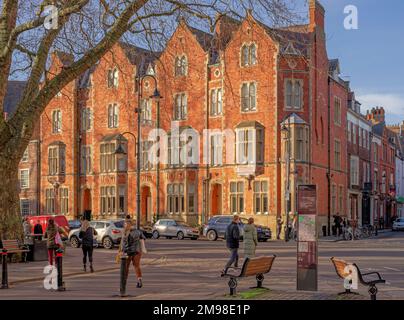 Straßenkreuzung mit einem alten Backsteingebäude, vorbei an einem überirdischen Baum. Bänke und eine Karte befinden sich im Vordergrund und die Leute sind auf der Straße. Stockfoto