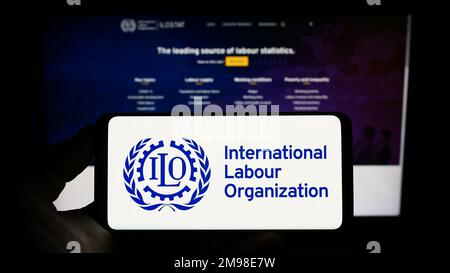 Person, die ein Smartphone mit dem Logo der Internationalen Arbeitsorganisation der Vereinten Nationen (IAO) auf dem Bildschirm vor der Website hält. Konzentrieren Sie sich auf das Display des Telefons. Stockfoto