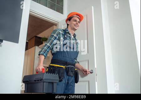 Kleiner Winkel eines freudigen Reparaturers mit dem Werkzeugkasten, der die Zimmertür in der Kundenwohnung öffnet Stockfoto