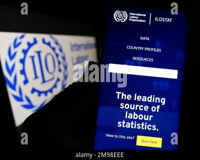 Person, die ein Mobiltelefon mit einer Webseite der Internationalen Arbeitsorganisation der Vereinten Nationen (IAO) auf dem Bildschirm mit Logo hält. Konzentrieren Sie sich auf die Mitte des Telefondisplays. Stockfoto