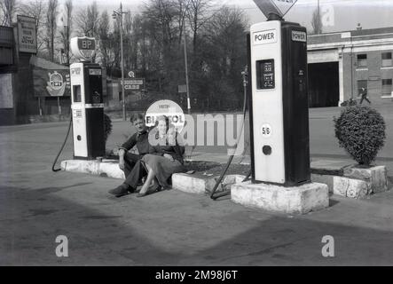 1950er, historisch, ein männlicher Automechaniker und weibliche Mitarbeiterin der Hargood Motor Co, die auf dem Vorplatz der Garage, Wilmslow Rd, Parrs Wood, Didsbury, Manchester, für ein Foto von Benzinpumpen für Power und BP-Treibstoff sitzt. England, Großbritannien. Der Preis für Power-Benzin liegt bei 5/6d (5s 6d) pro Gallone, teuer, da es die Zeit der Suez-Krise war. Auf der anderen Straßenseite befindet sich ein großer Busbahnhof, der die Stadt Manchester bedient. Am Eingang zur Eisenbahnbrücke, in der Nähe des Bahnhofs East Didsbury & Parrs Wood, befindet sich ein Schild für Morris Cars. Stockfoto