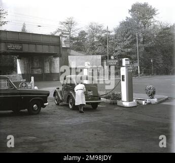 Ende 1950er, historisch, eine weibliche Gargenbegleiterin, die Benzin in einem beliebten Ford-Auto in der Hargood Motor Co an der Wilmslow Rd, Parrs Wood, Manchester, England, Großbritannien, betreibt. Der Treibstoff ist Regent, eine Marke der Regent Oil Company, die 1947 von Texaco of America und Trindad leaseholds in Großbritannien gegründet wurde. Hinter der Garage, eine Eisenbahnbrücke, Schild für den Bahnhof mit der Aufschrift „British Railways“, East Didbsbury & Parrs Wood“, Stockfoto