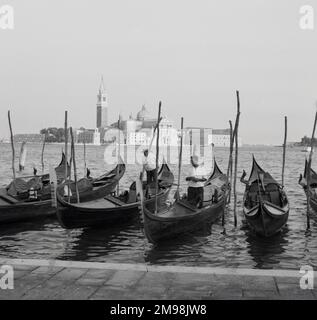 1950er, historische, traditionelle venezianische Ruderboote oder Gondeln in der Lagune von Venedig, einer geschlossenen Bucht an der Adria, Italien. Zwei Gondoliere stehen in ihren Booten. Die flachen Boote werden für Fahrten rund um die Inseln von Venedig verwendet, ein Flickwerk aus vielen Inseln, wobei die berühmte historische Stadt Venedig in der Ferne zu sehen ist. Die Hauptstadt der Region Venetien wurde 1866 Teil von Italien. Stockfoto