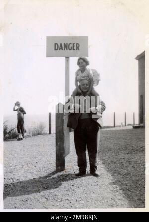 Ein humorvolles Foto eines jungen Mädchens, das auf den Schultern seines Großvaters sitzt, steht neben einem „Danger“-Schild am Rand einer Klippe (wahrscheinlich in einem britischen Küstenresort). Stockfoto