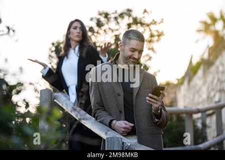 Ein Mann, der auf sein Handy schaut, mit einer wütenden Freundin dahinter Stockfoto