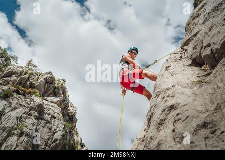 Aktiver Bergsteiger Mann mittleren Alters in Schutzhelm Blick auf Kamera, während Abseilen von Felswand mit Seil mit Sicherungsvorrichtung und Klettern ha Stockfoto