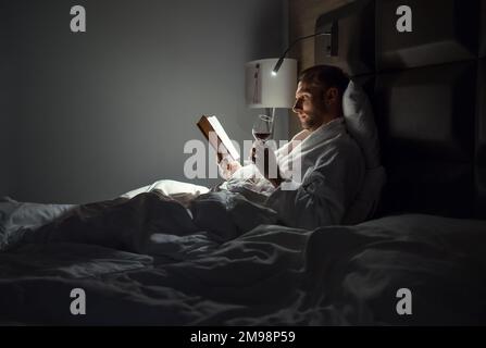 Ein Mann mittleren Alters, der sich im Bett entspannt und ein Buch mit einem Glas Rotwein hält, wobei die Nachttischlampe eingeschaltet ist. Abendliche Entspannung, Hobbys, Freizeit-Konzept. Stockfoto