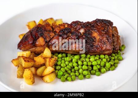 Gebratener Schweinebauch mit knuspriger Haut und Kartoffeln und Erbsen auf weißem Teller, Nahaufnahme. Stockfoto