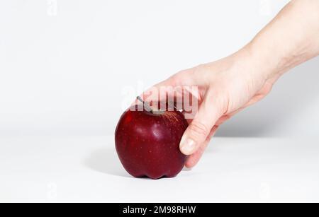 Roter reifer Apfel in der Hand eines Mannes, auf weißem Hintergrund mit hartem Schatten. Die Hand nimmt den Apfel auf den Tisch in der Küche. Der Begriff der Heilung Stockfoto