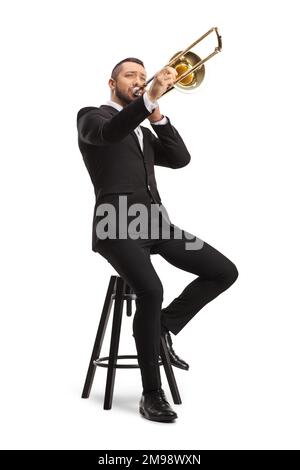 Männlicher Musiker, der auf einem Stuhl sitzt und eine Posaune spielt, isoliert auf weißem Hintergrund Stockfoto