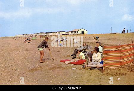 Holiday Beach Szene - Heacham, Norfolk - alle wesentlichen Merkmale des britischen Strandurlaubs sind hier - mehrere Schichten Kleidung, ein gestreifter Windschutz, Opa nickt in einem ausklappbaren Stuhl, ein hellroter Lolly, der widerwillig verzehrt wird, Schuhe, die getragen wurden, um dem Unbehagen beim Gehen auf Kieseln entgegenzuwirken, und eine Reihe von verschlossenen Strandhütten, die sich bis in die Unendlichkeit wiederholten Stockfoto