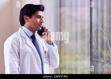 Männlicher Arzt Trägt Weißen Mantel Steht Im Krankenhaus Korridor Und Spricht Auf Dem Handy Stockfoto