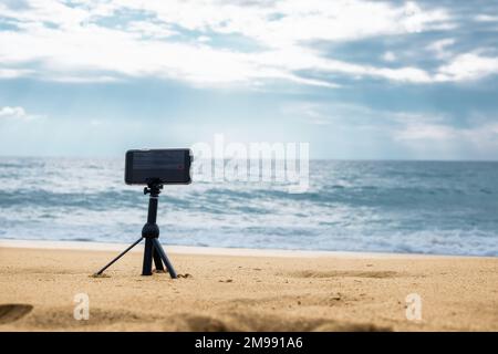 Eine Smartphone-Kamera auf einem Stativ im Vordergrund steht an der Küste Stockfoto