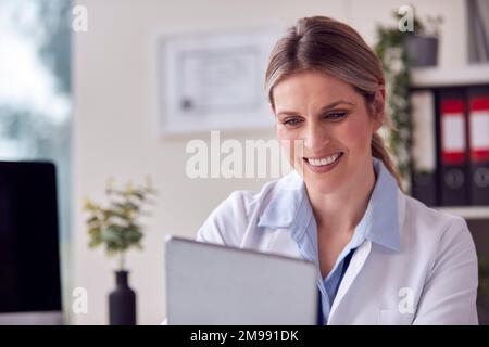 Ärztin oder Hausärztin mit weißem Mantel, die mit einem Digital Tablet am Schreibtisch sitzt Stockfoto