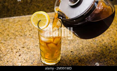 Frisch gebrühten Eistee in klares Glas gießen. Ein Krug Tee, der in ein Glas Eis mit einer Zitronenscheibe gießt. Stockfoto