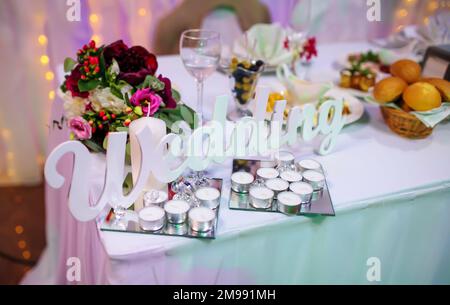 Wort Hochzeit, weiße Holzbriefe mit Kerzen und Blumen auf dem Feiertagstisch. Hochzeitsdekor. Das Hochzeitszeremoniekonzept feiern Stockfoto