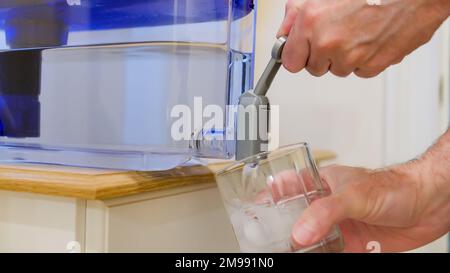 Abgabe von frischem gefiltertem Wasser aus dem Krug zur Wasseraufbereitung zu Hause. Mann gießt sauberes Wasser in ein Glas mit Eis aus dem Wasserausgießer des Behälters. Stockfoto