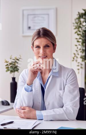Porträt einer lächelnden Ärztin oder Hausärztin mit weißem Mantel im Büro, die am Schreibtisch sitzt Stockfoto