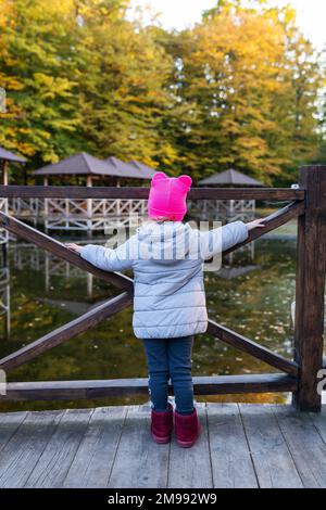 Ein kleines Mädchen mit rosa Hut steht mit dem Rücken neben dem Teich und genießt einen warmen Herbsttag Stockfoto