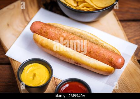 Klassischer Fast-Food-Hotdog mit Senf auf einem rustikalen Holzbrett. Serviert mit Kartoffelchips und Soßen. Stockfoto