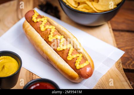 Klassischer Fast-Food-Hotdog mit Senf auf einem rustikalen Holzbrett. Serviert mit Kartoffelchips und Soßen. Stockfoto