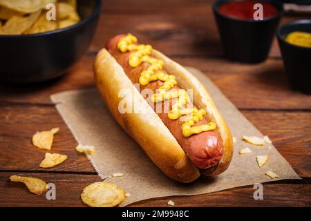 Klassischer Fast-Food-Hotdog mit Senf auf einem rustikalen Holzbrett. Serviert mit Kartoffelchips und Soßen. Stockfoto