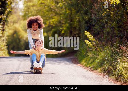 Kinder, Die Spaß Haben Mit Dem Jungen, Das Mädchen Auf Dem Skateboard Auf Der Country Road Schiebt Stockfoto