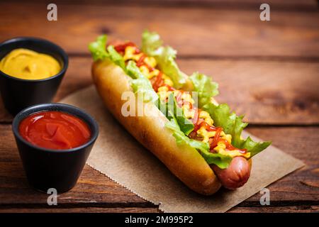 Hot Dog mit Senf, Ketchup und grünem Salat im Brötchen, serviert mit Kartoffelchips auf einem rustikalen Holzbrett. Fast Food und köstlich. Stockfoto