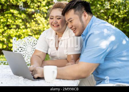 Ein junges asiatisches, dickes Paar, das im grünen Park im Freien mit einem Laptop unterwegs ist. Glücklicher lächelnder Mann und Frau, die Remote arbeiten und ein eigenes Unternehmen gründen Stockfoto
