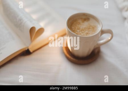 Kuscheliges Morgenleben mit einer Kaffeetasse, Nadeln und einem Buch mit Seiten, die im Bett in Herzform gefaltet sind Stockfoto