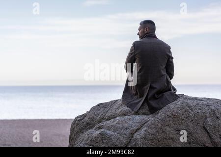 Ein Mann sitzt auf einem Felsen am Strand und schaut aufs Meer Stockfoto