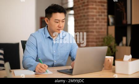 Selbstbewusster asiatischer Mann mittleren Alters, der Notizen macht, an einem Notebook im Heimbüro arbeitet, Panorama, freier Platz Stockfoto