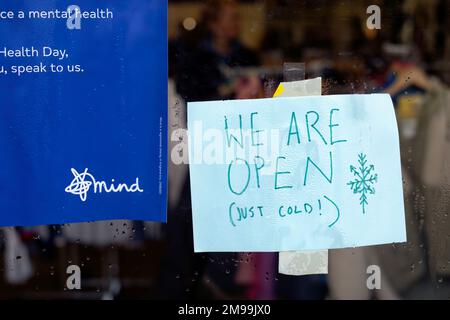 Ein amüsantes Schild an der Tür eines Wohltätigkeitsladens. Auf dem Schild steht, dass der Laden geöffnet ist, aber dass das Personal kalt ist.die Energiekostenkrise in Großbritannien beeinträchtigt das Geschäft Stockfoto