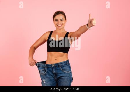 Erfolgreiche Diät. Happy Slim Woman Trägt Übergroße Jeans Und Zeigt Daumen Hoch Stockfoto