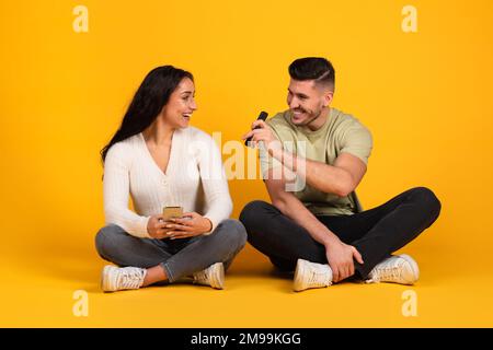 Ein lächelnder arabischer Mann, der seiner Frau das Smartphone zeigt, eine App empfiehlt, auf dem Boden sitzt Stockfoto