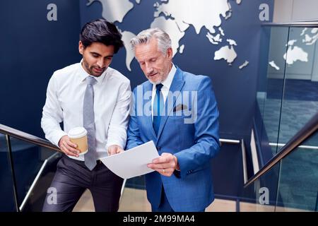 Männliche Kollegen Treffen Sich Auf Der Treppe Des Büros Und Diskutieren Das Dokument Mit Der Weltkarte Im Hintergrund Stockfoto