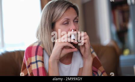 Eine erkältete Frau hält ein Nasenspray in der Hand, während sie zu Hause auf der Couch sitzt. Stockfoto