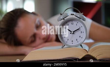 Müde Frau schläft auf einem Tisch mit Buch und Wecker. Stockfoto