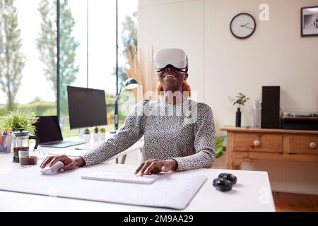 Frau, die vom Home Office aus am Schreibtisch sitzt und ein VR-Headset mit AR-Technologie trägt Stockfoto