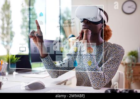 Frau, die auf den projizierten Bildschirm für Smart Home schaut, sitzt am Schreibtisch und trägt ein VR-Headset Stockfoto