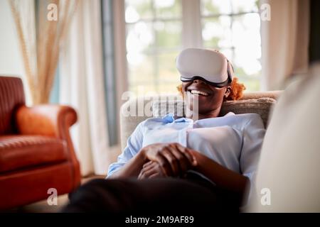 Lachende Frau, die zu Hause auf dem Sofa liegt, ein VR-Headset trägt und mit AR-Technologie interagiert Stockfoto