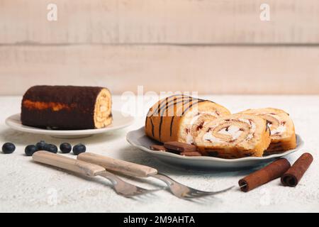 Teller mit leckeren Kuchenrollen, Zimt, Kaffeebohnen und Gabeln auf leichtem Grunge-Hintergrund Stockfoto