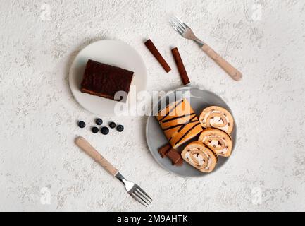 Teller mit leckeren Kuchenrollen, Zimt, Kaffeebohnen, Schokolade und Gabeln auf leichtem Grunge-Hintergrund Stockfoto