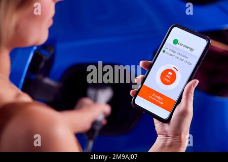 Frau Lädt Elektroauto Mit Kabel Auf, Indem Sie Die App Auf Dem Telefon Verwendet, Um Den Akkustand Zu Überwachen Stockfoto