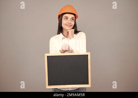 Junge lustige Frau Bauleiterin. Frau Baumeister isoliertes Porträt mit Schutzhelm und Brett. Grauer Hintergrund, leerer Speicherplatz, Kopierbereich. Stockfoto