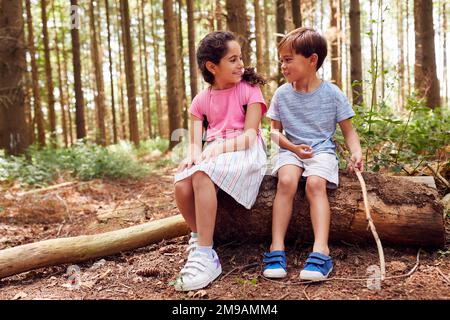 Smiling Boy And Girl Walking In Summer Woodland Sitting On Log Mit Rucksäcken Stockfoto