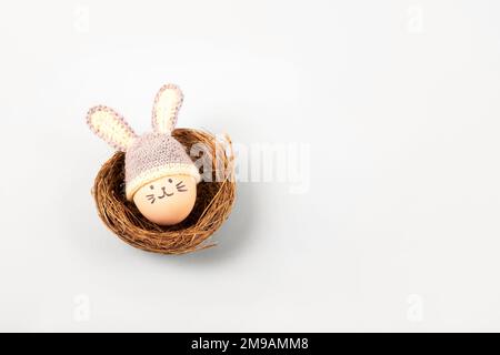 Osterei mit lustigem Gesicht und grauer Häkelmütze mit Hasenohren in einem dekorativen Sisalnest auf grauem Hintergrund. Frohes Osterkonzept. Begrüßung C. Stockfoto