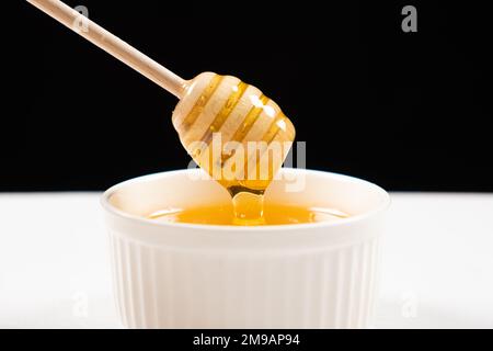 Honig in einer weißen Keramikschüssel mit einem Holzlöffel auf schwarzem Hintergrund. Stockfoto