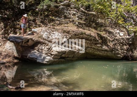 Eine junge, gut aussehende Frau, die an der felsigen Klippe über dem kleinen grünen See inmitten unberührter Natur steht. Der See ist von Wald umgeben Stockfoto