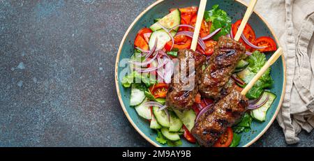 Gegrillte Spießfleisch-Rindfleisch-Kebabs auf Stöcken serviert mit frischem Gemüsesalat auf einem Teller auf rustikalem Betonhintergrund von oben. Traditioneller Mittelteil Stockfoto